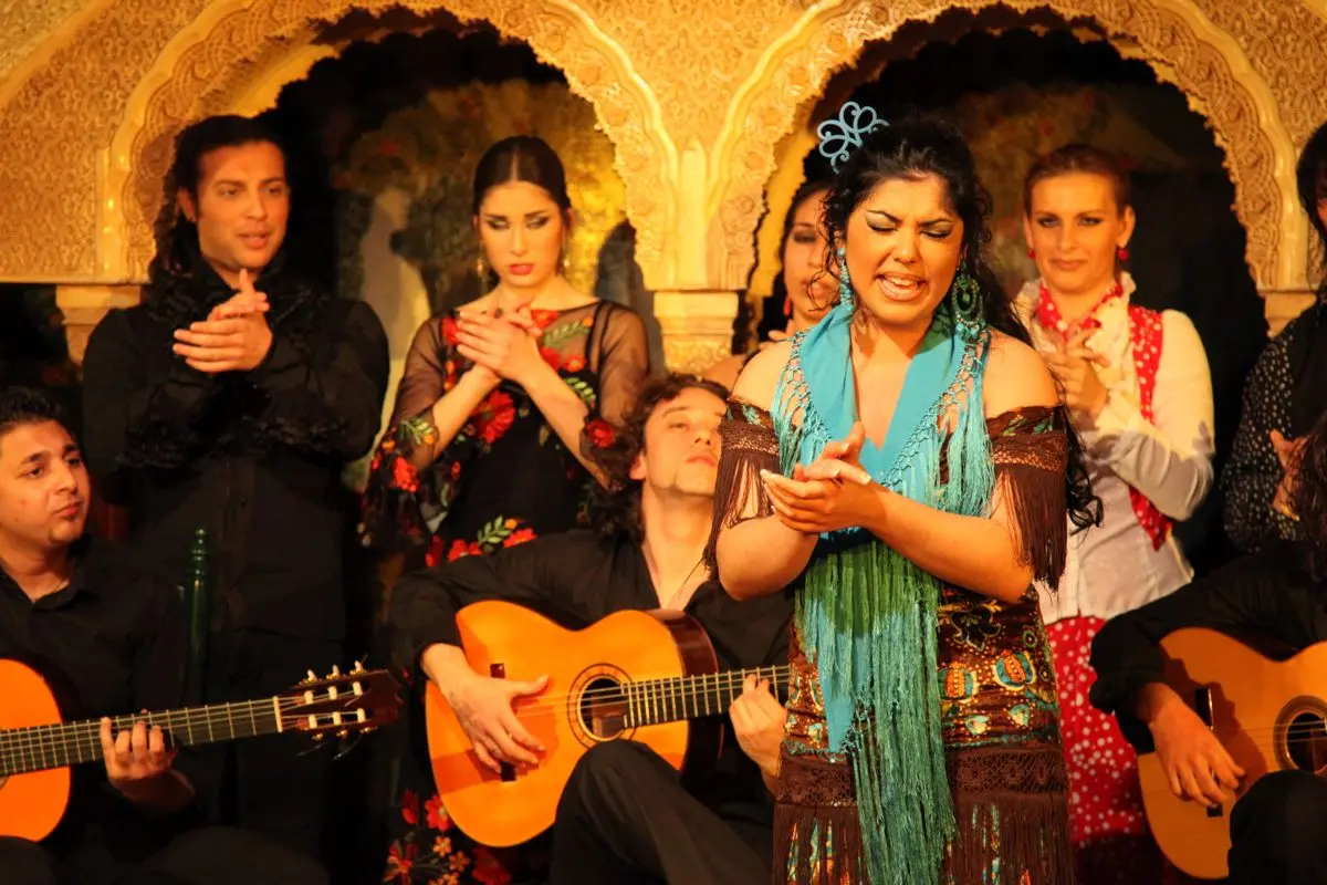 A flamenco singer clapping in a turquoise shawl, with guitarists and dancers behind her on a tablao stage